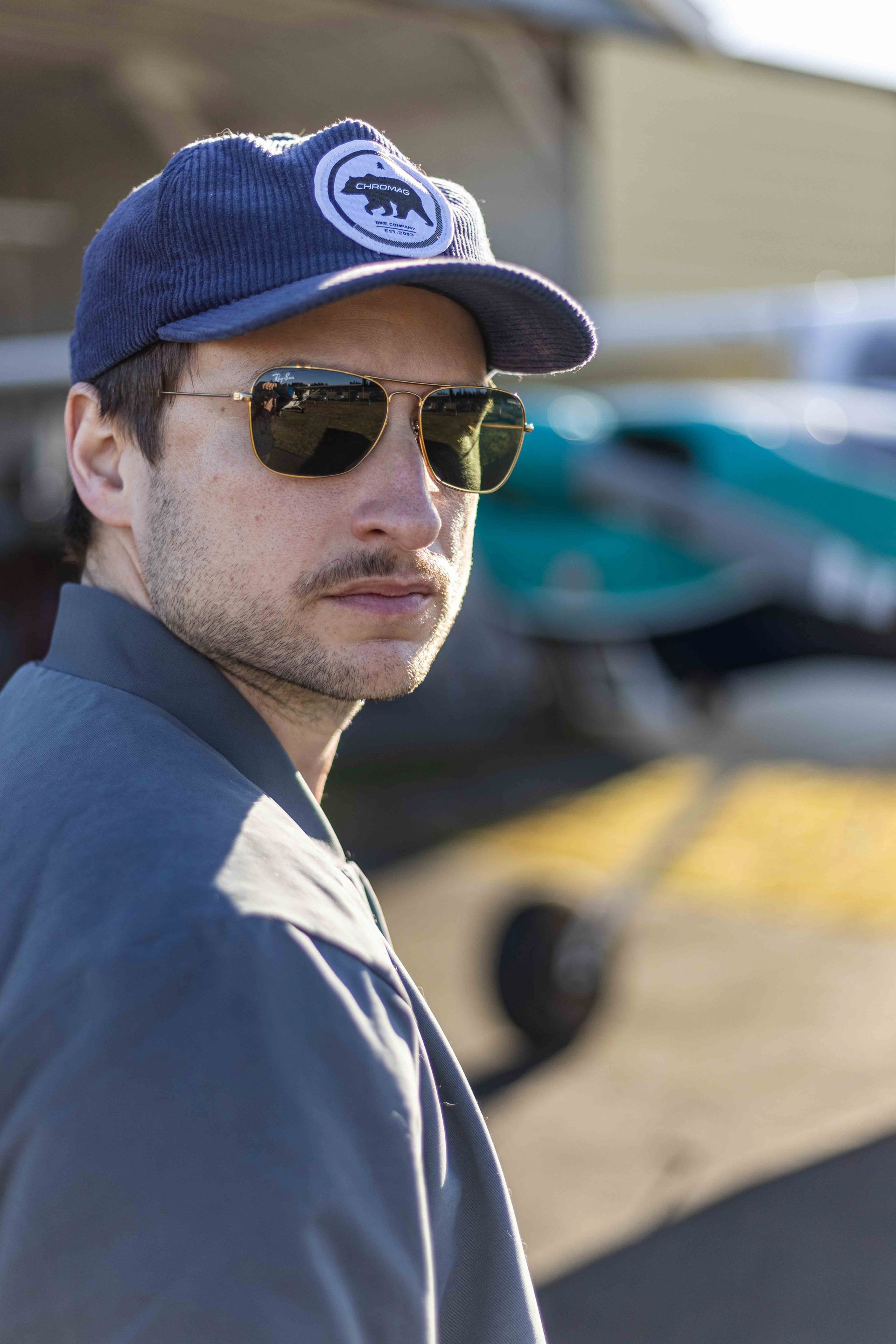 Man in sunglasses and cap with a bear logo, near an airplane, looking over shoulder.