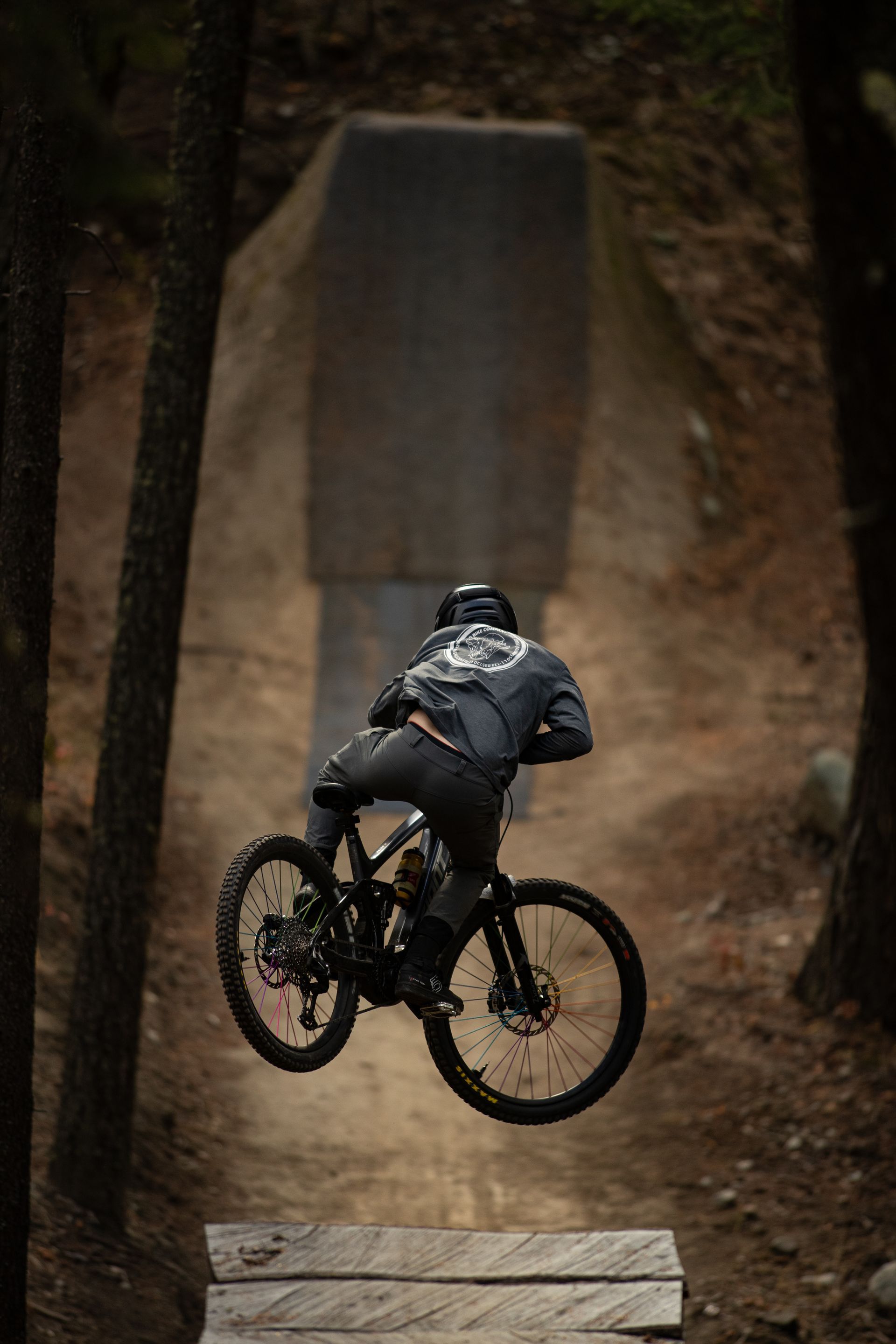 Mountain biker in mid-air above wooden ramp in a forest, dark clothing, bike details visible.