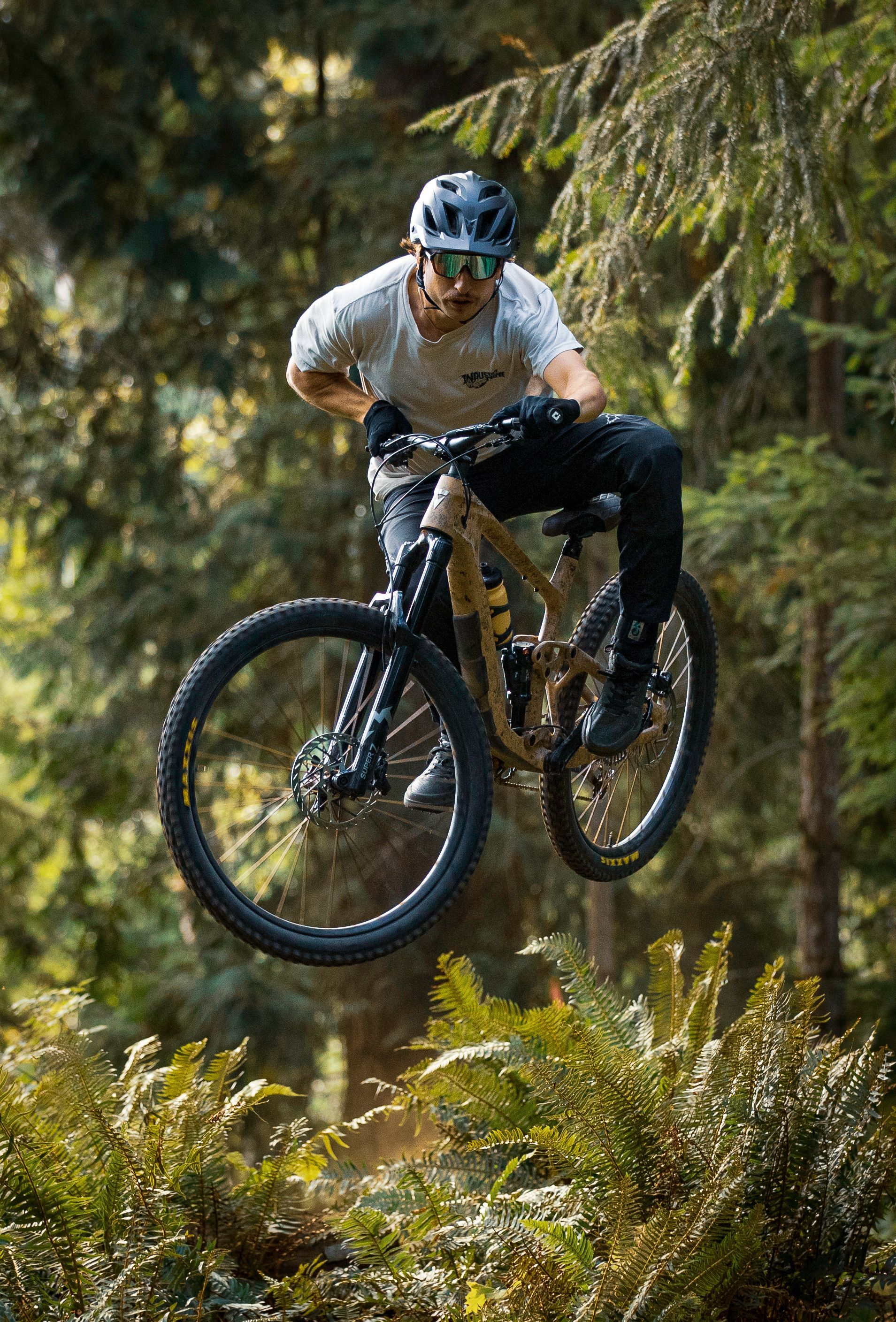Mountain biker in mid-air over greenery, wearing a helmet, outdoors in forest.