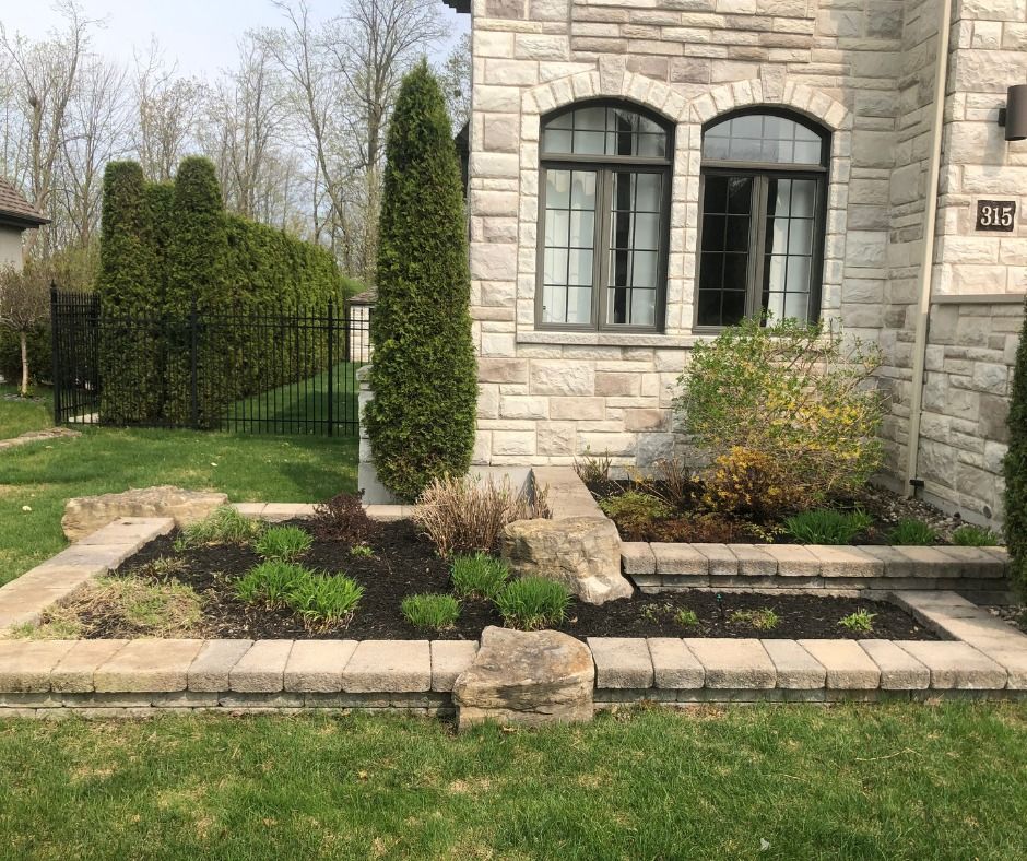 Façade en pierre d'une maison avec fenêtres cintrées et petit jardin paysager bordant une pelouse.