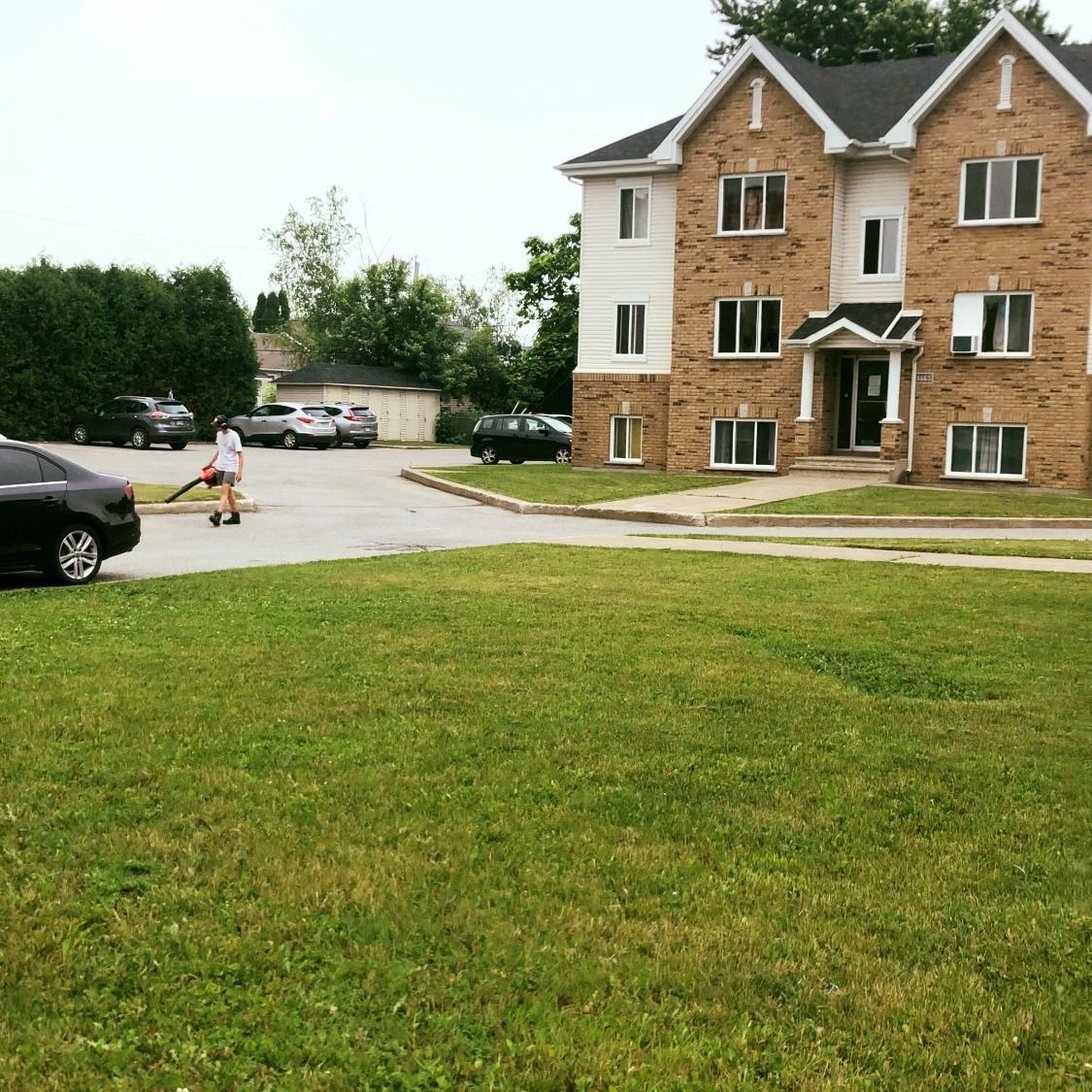 Immeuble d'appartements de banlieue avec une pelouse verte, des voitures garées et un enfant sur une trottinette près de l'allée.