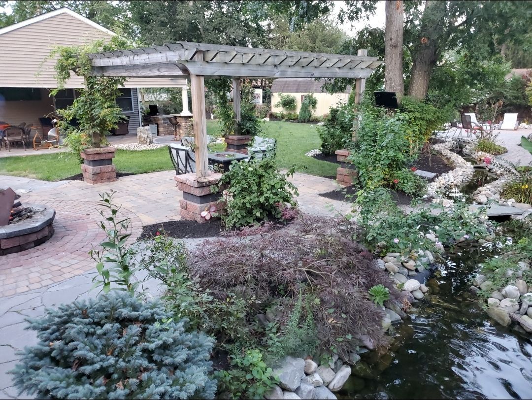 Backyard landscape with pergola, pond, brick patio, and lush greenery.