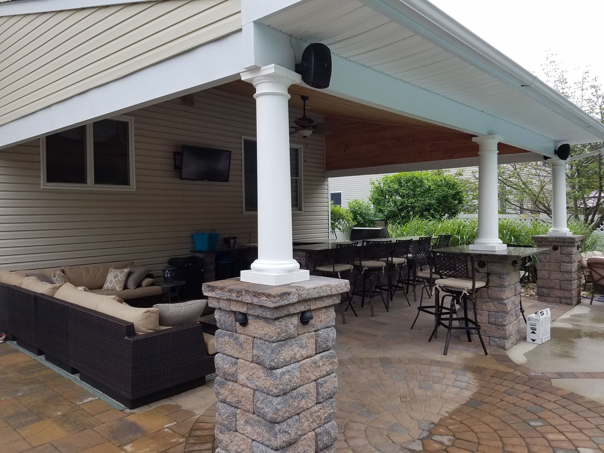 Covered patio with seating, bar, and television; tan siding, brick columns, and brown furniture.
