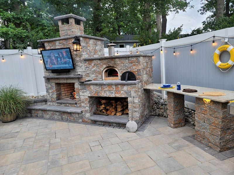 Stone outdoor kitchen with pizza oven, fireplace, and counter under a TV, with lights strung on a fence.