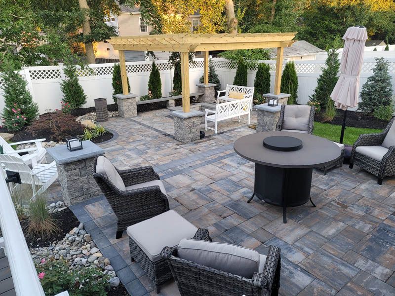Patio with outdoor furniture, pergola, and fire pit. Paved with multi-color stone, with white fence in background.