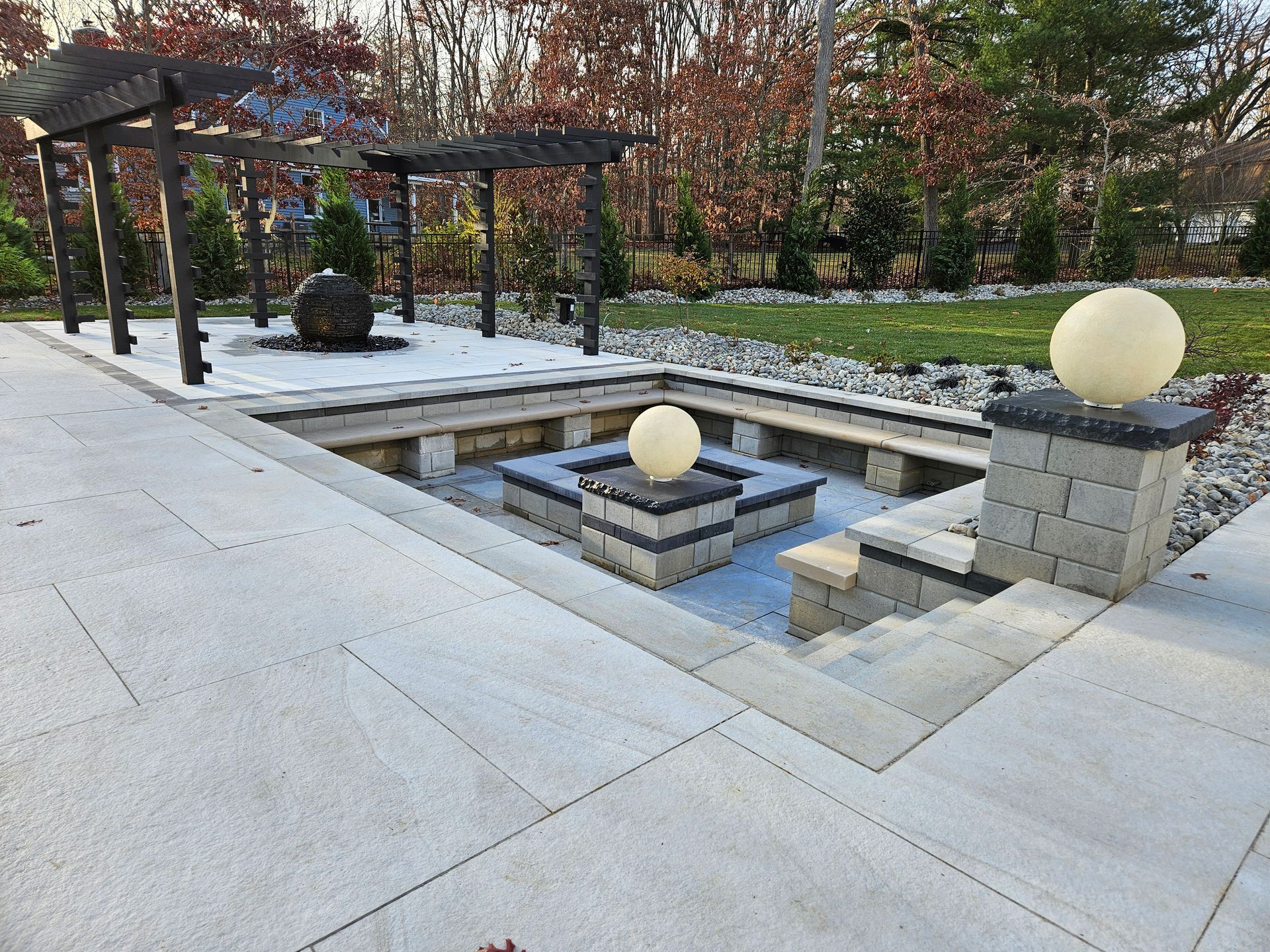 A modern sunken patio with a square fire pit, stone seating, spherical light fixtures, and a pergola in a wooded yard.