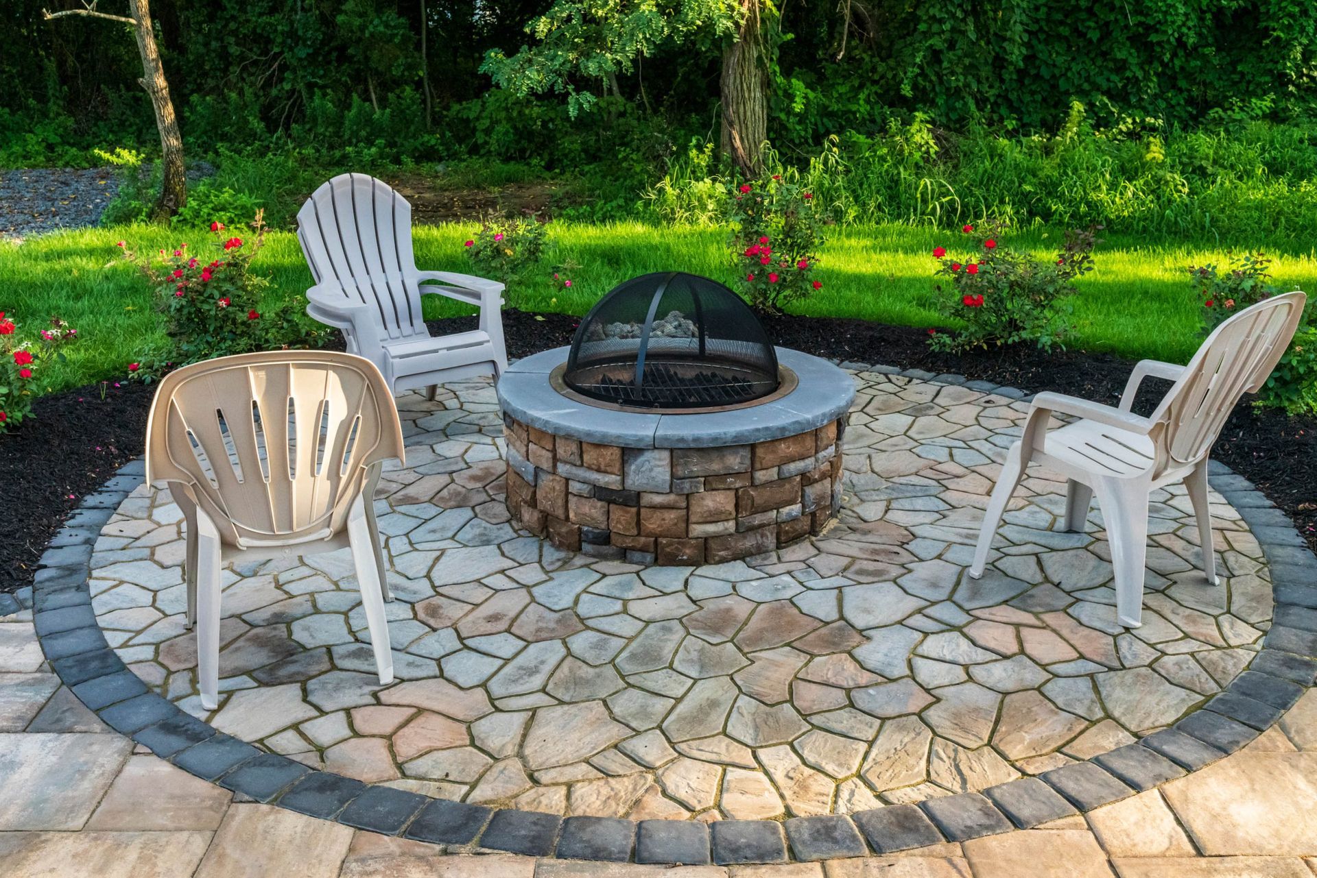 Fire pit surrounded by stone patio with three plastic chairs in a backyard setting.