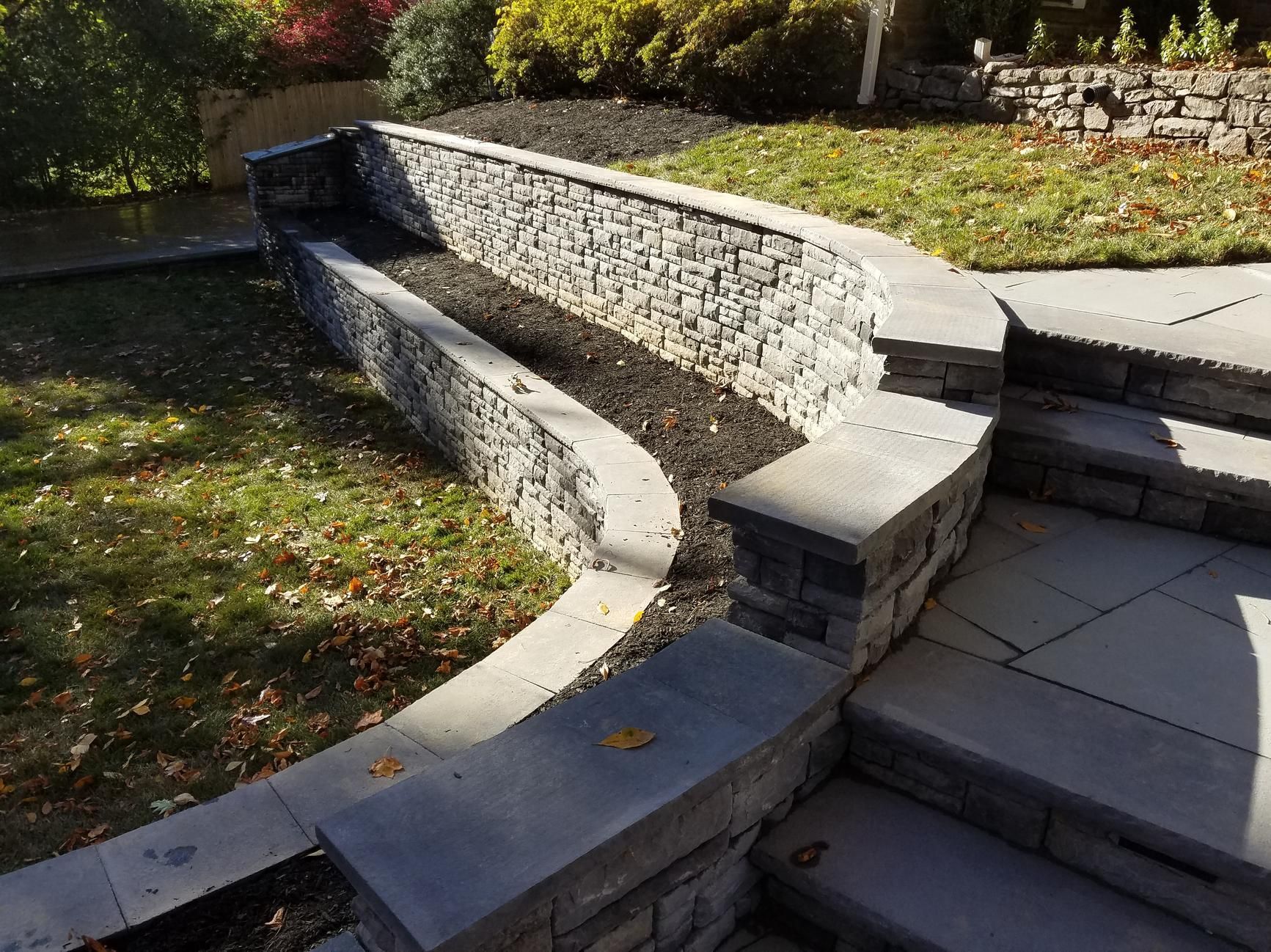 Stone steps leading up to a curved stone retaining wall with a planter bed; grassy area.