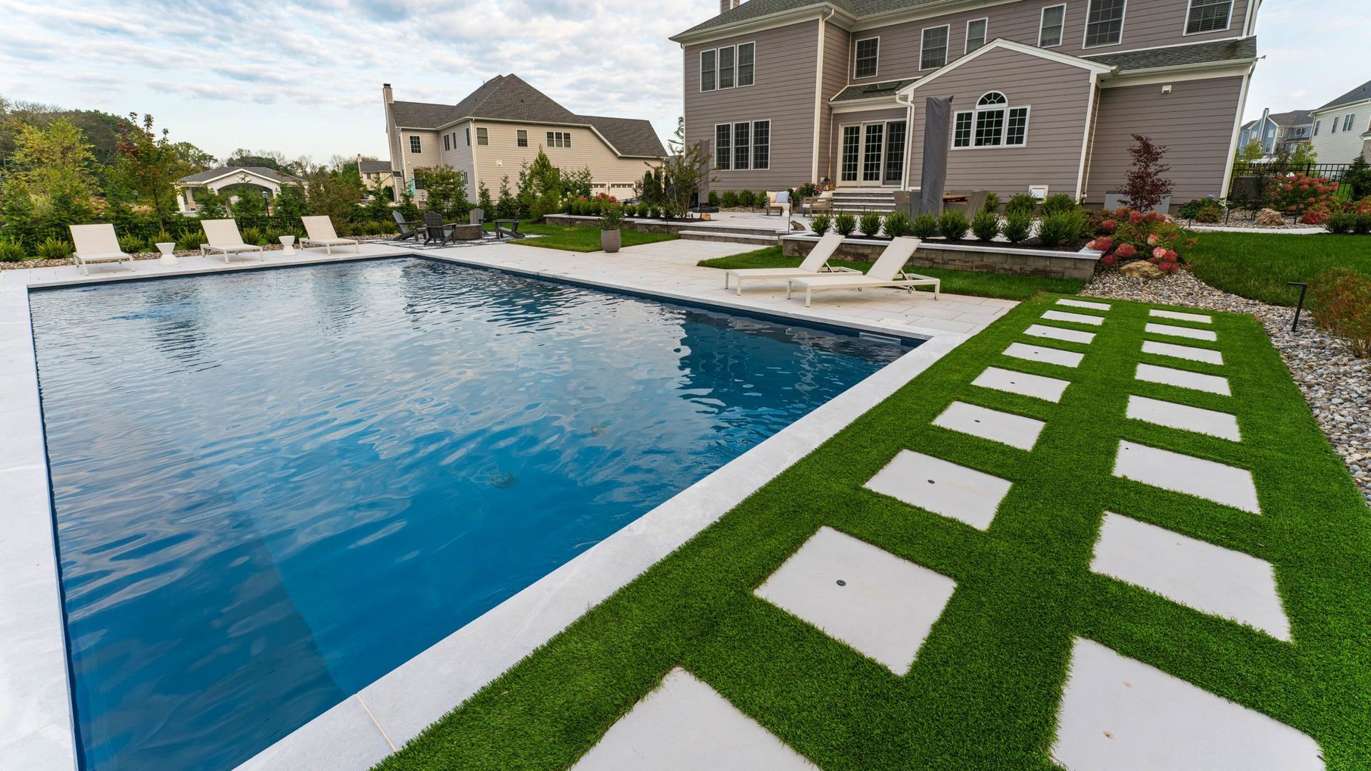 Backyard pool with white concrete border, green lawn, and stone path leading to a multi-story home.