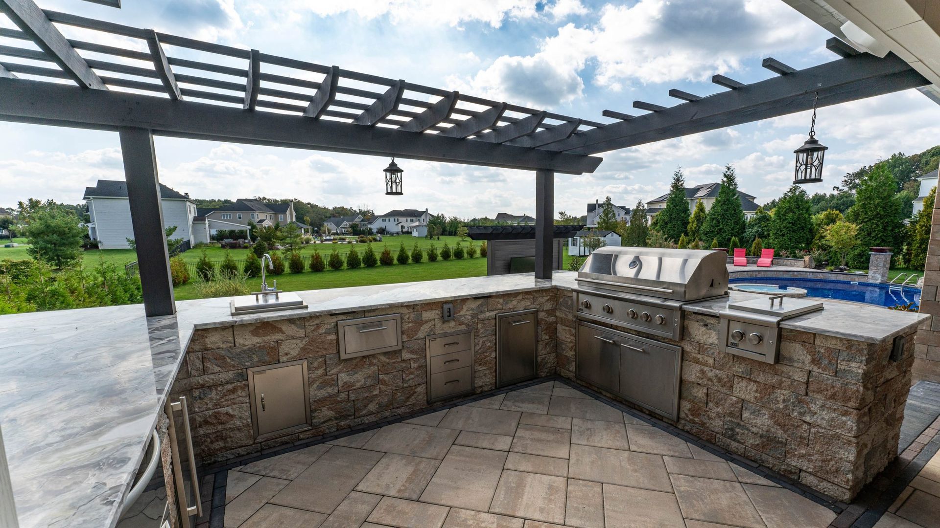 Outdoor kitchen under a pergola with built-in grill, sink, and stone counter, overlooking a lawn.