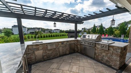 Outdoor kitchen with grill, sink, stone facade, pergola, and view of landscape under a cloudy sky.