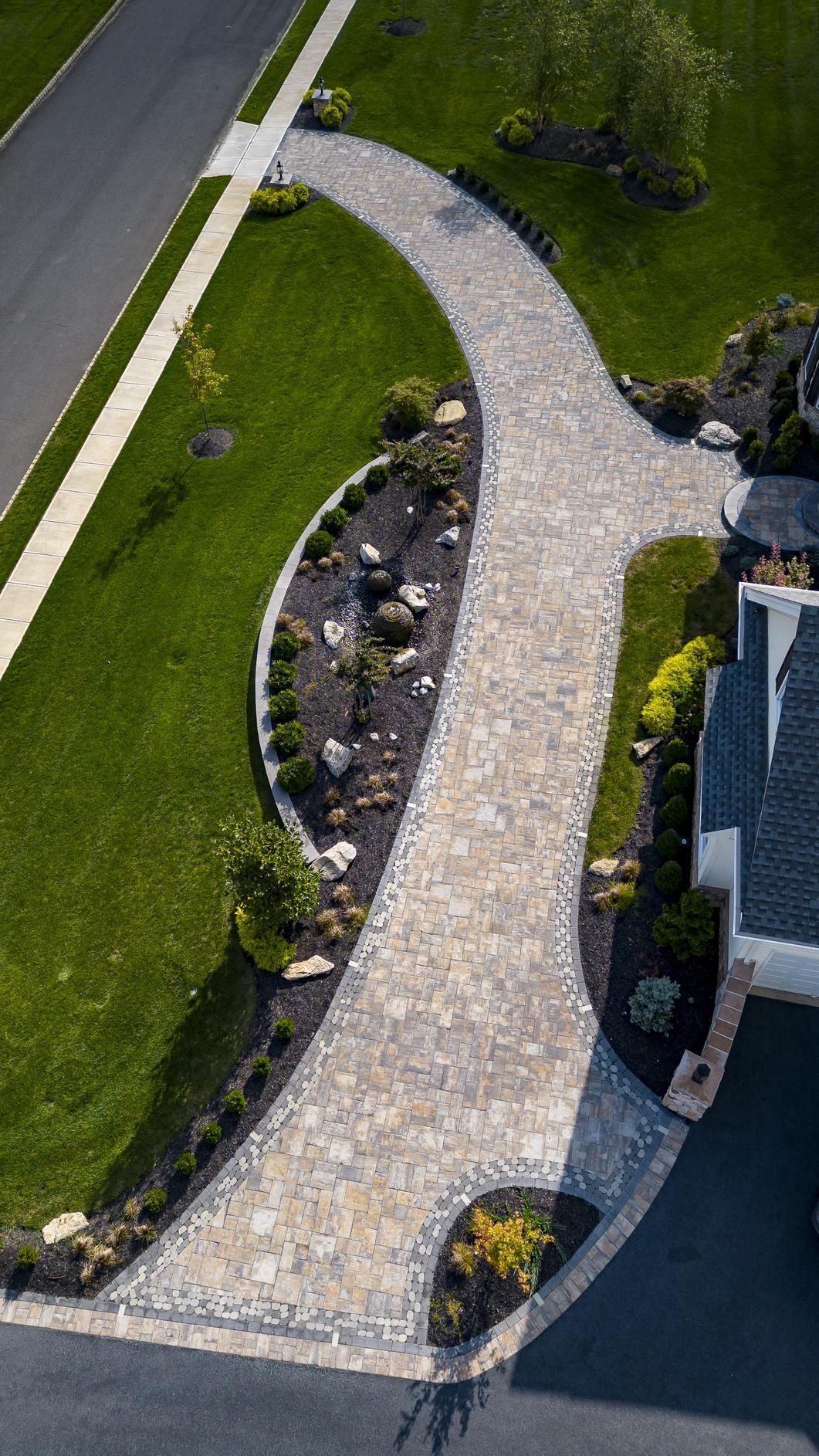 Stone paver driveway curves past landscaped beds, green lawn.