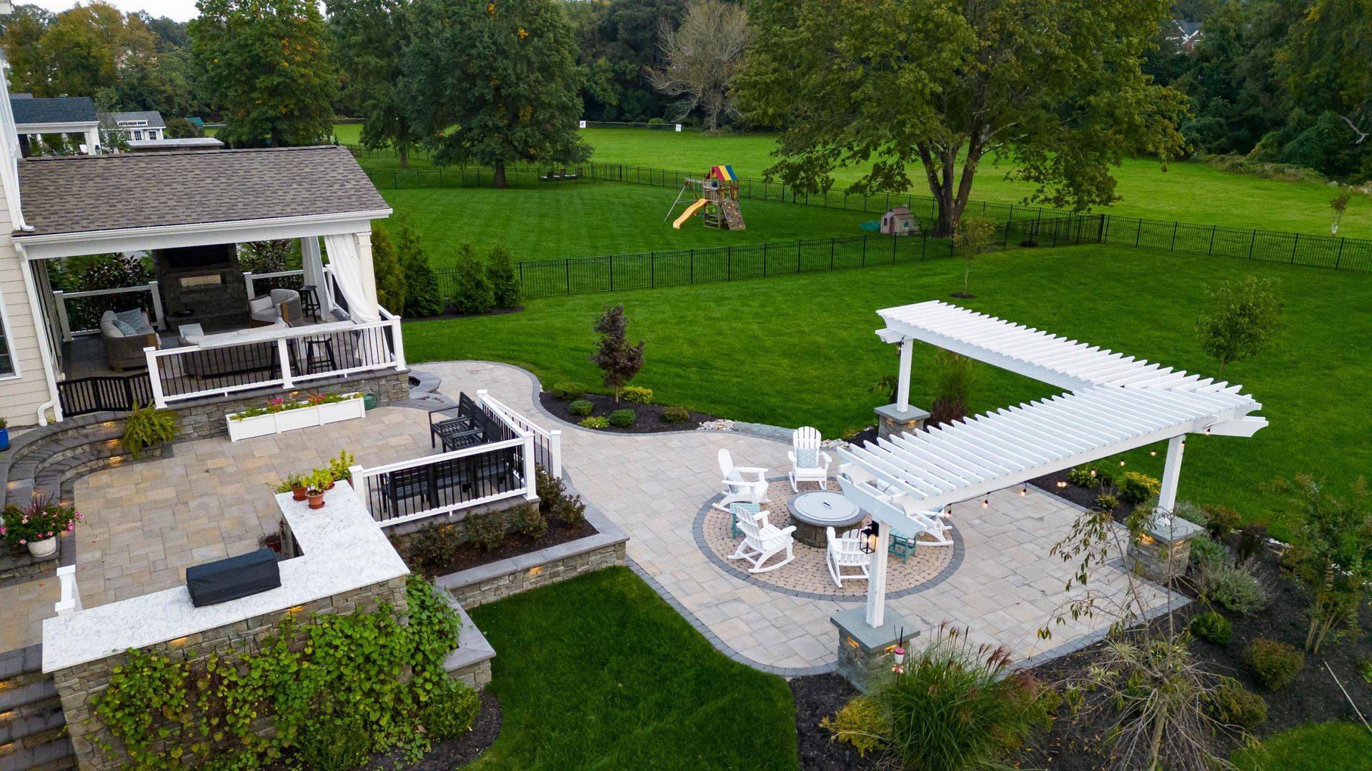 Patio with white pergola and seating, adjacent to a house with a covered porch and expansive green lawn.