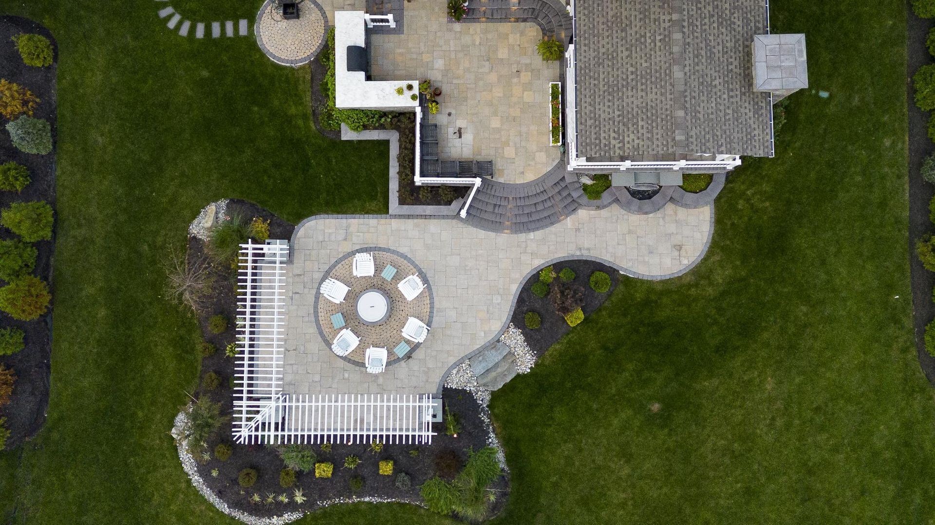 Overhead view of a backyard patio with stone pavers, seating, and a pergola, surrounded by green grass.