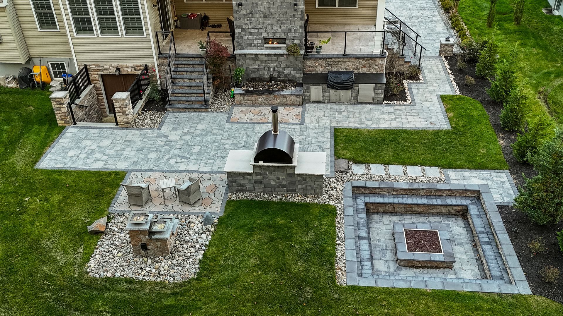 Aerial view of a backyard patio with a pizza oven, fire pit, seating areas, and manicured green lawn.