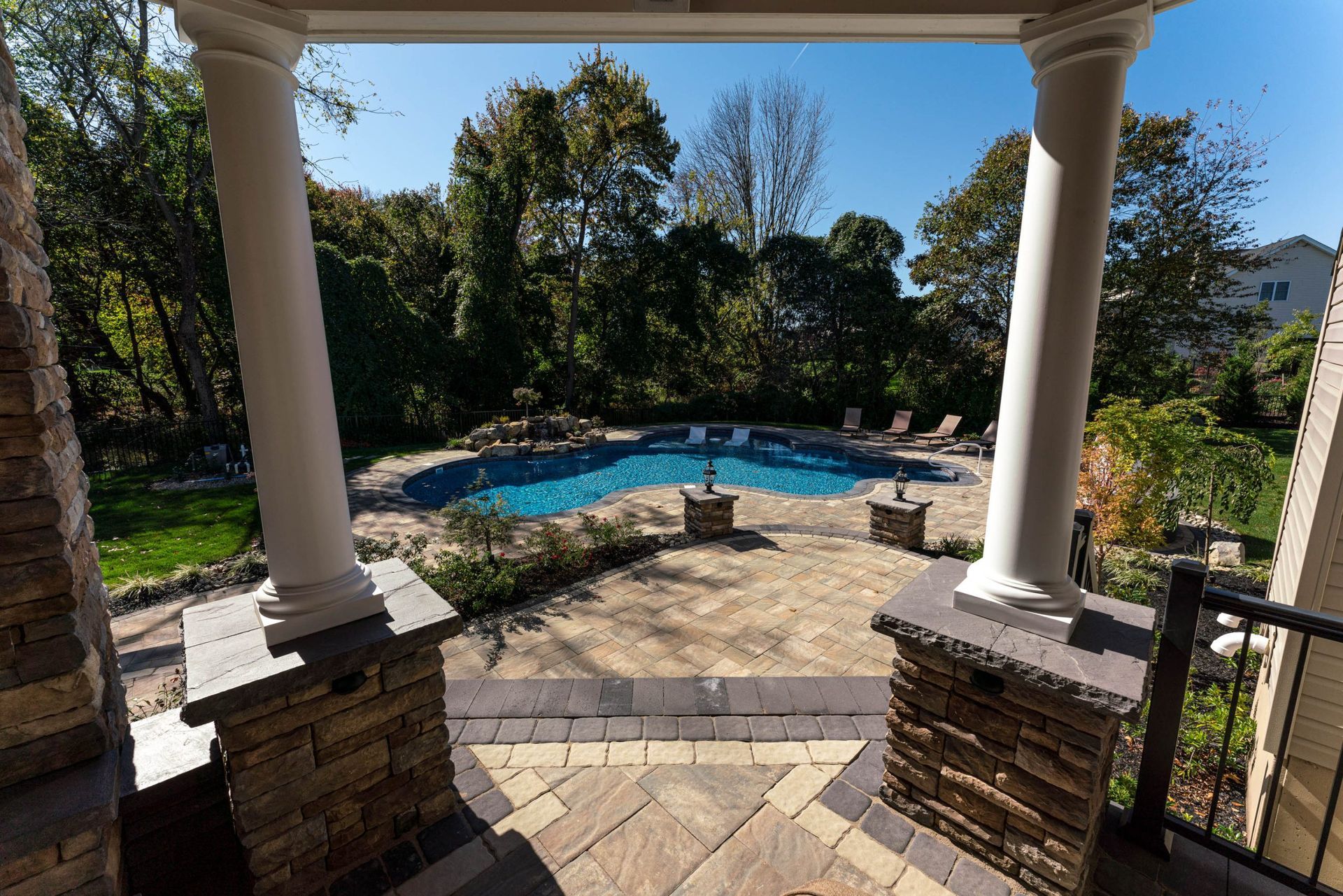 View of a pool through a porch. The patio has stone paving and pillars. Trees and blue sky are in the background.
