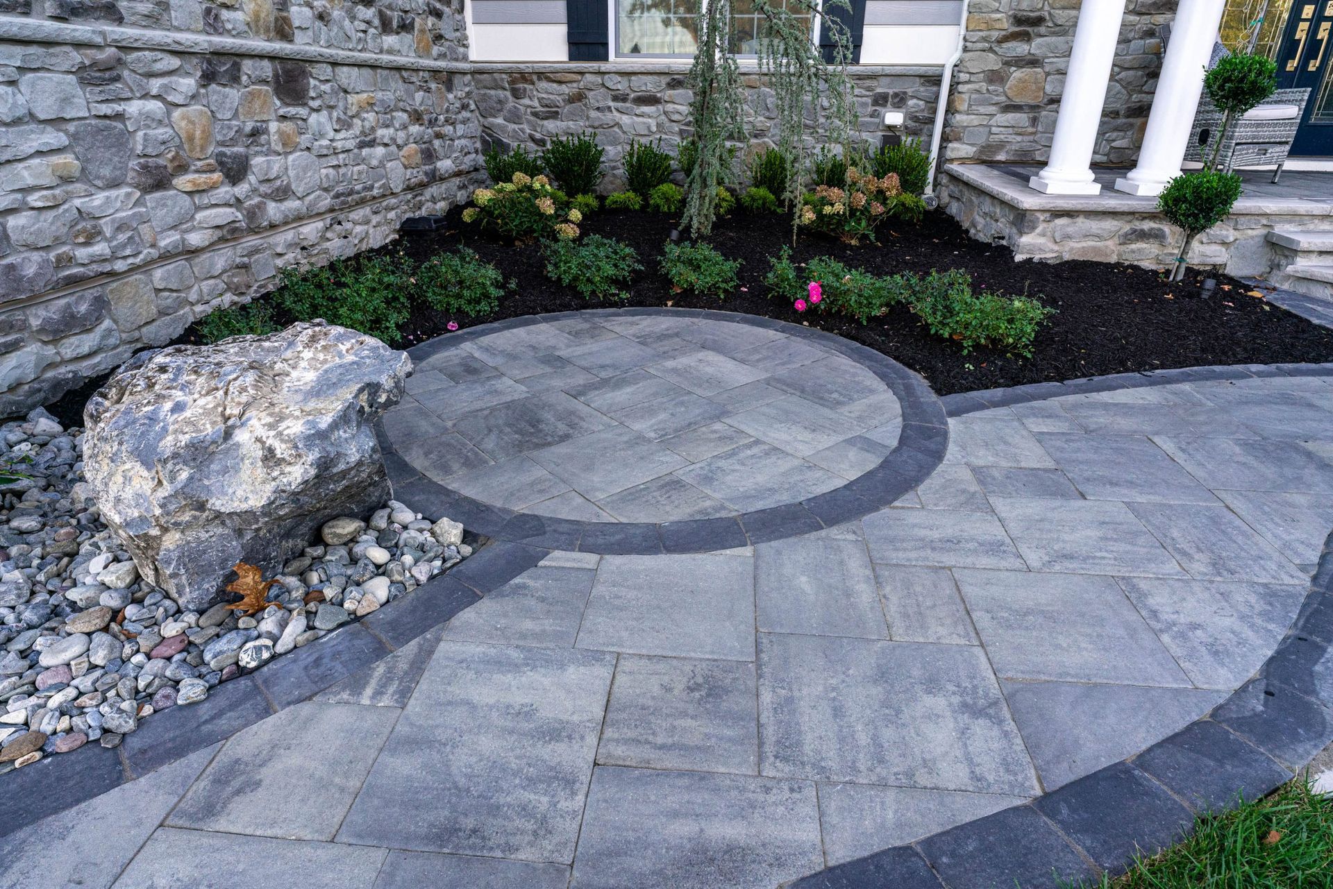 Stone patio with circular design, landscaping, and pathway in front of a building.