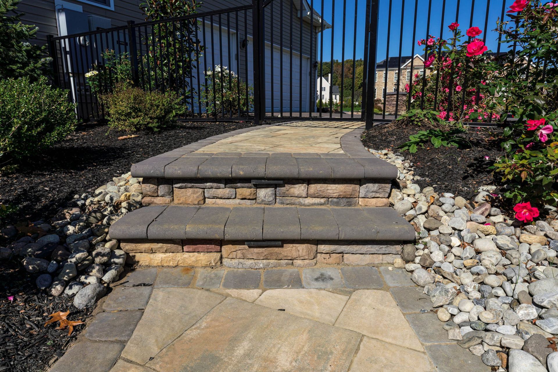 Stone steps leading to a black metal gate, bordered by flowers, rocks, and landscaping.