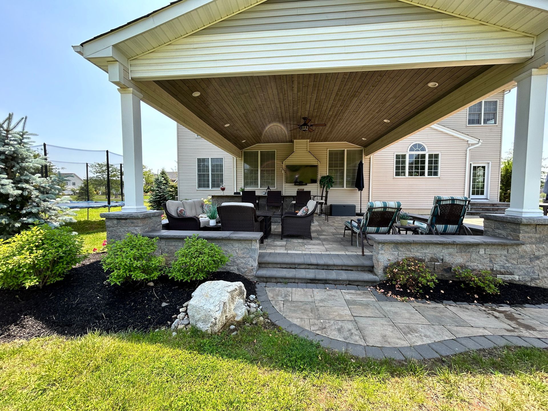A covered stone patio with outdoor seating, stairs, and a landscaped lawn area on a sunny day.