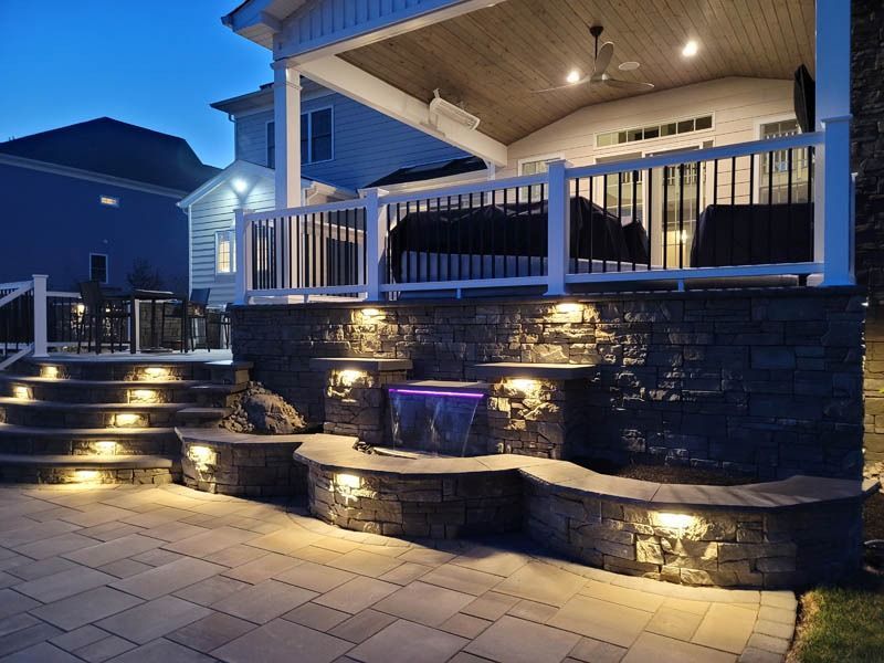 Night view of a multi-level outdoor patio with stone walls, stairs, and a waterfall feature, all illuminated.