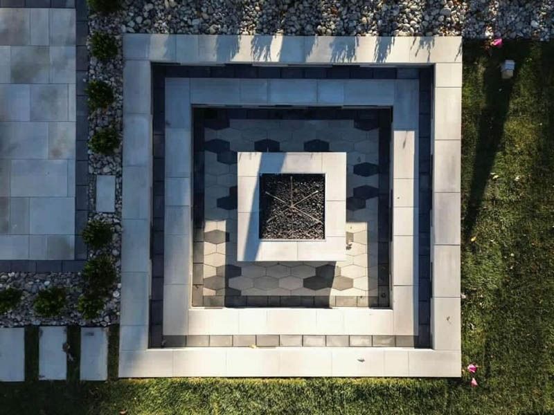 Overhead view of a square, tiered patio with a central fire pit, surrounded by gravel and grass.