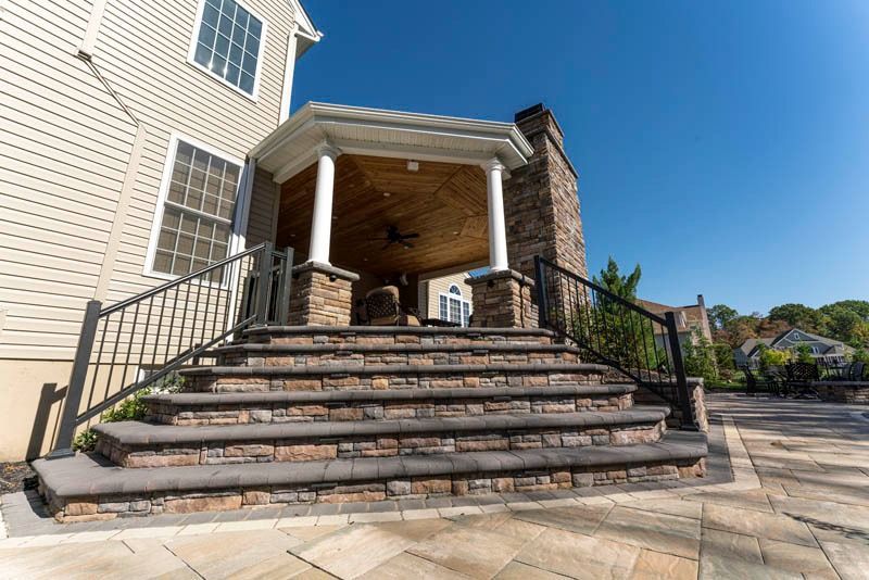 Outdoor stone steps leading to a covered patio with a brick chimney and metal railings against a blue sky.