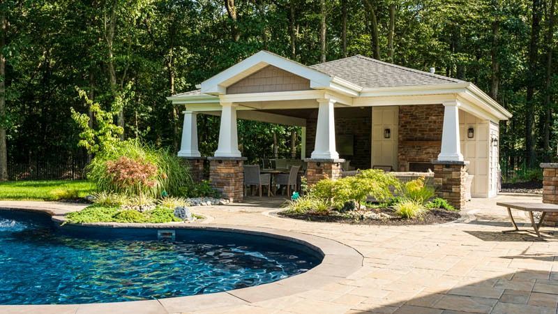 Poolside cabana with pillars, stone accents, and a backyard pool surrounded by lush landscaping.