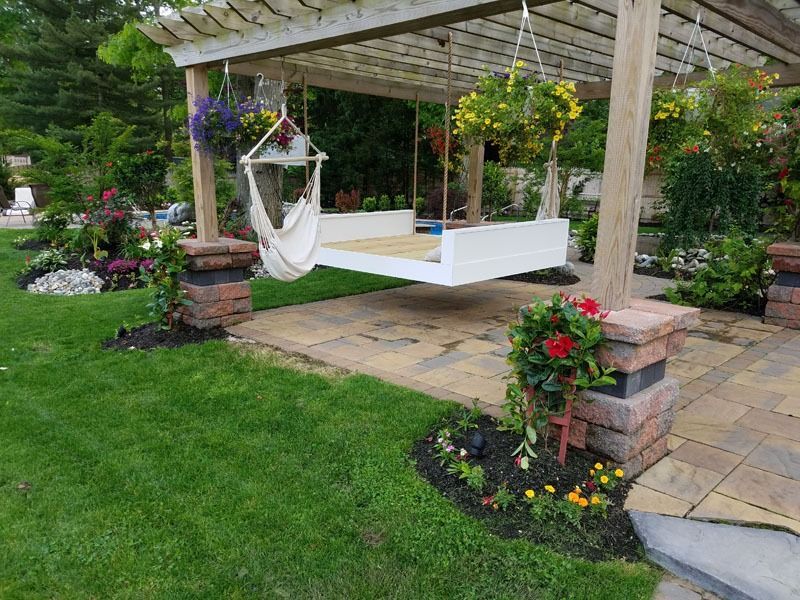 Pergola with hanging swing bed and hammock, surrounded by flower beds and lush green lawn.