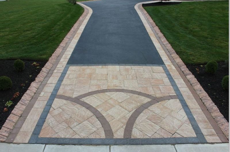 Driveway with brick-bordered asphalt path. Decorative stone section with curved pattern, surrounded by green lawn.