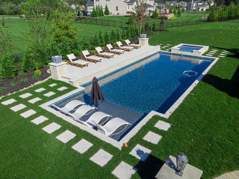Rectangular pool with lounge chairs, surrounded by grass and stone pavers in a backyard.