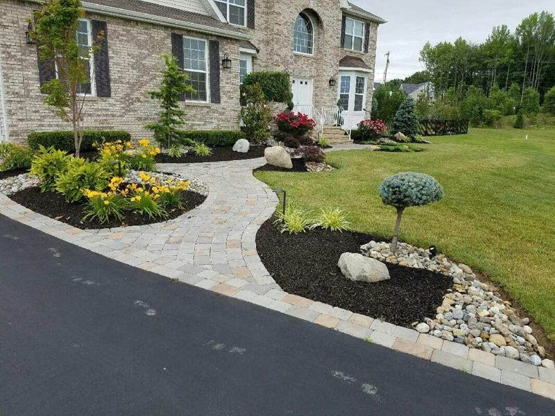 Stone walkway curves from driveway to a house with landscaping. Green grass, mulch, flowers, and trees are visible.