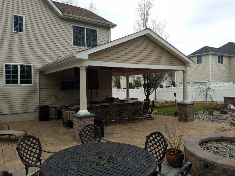 Backyard patio with covered bar, seating, and fire pit. Beige and brown tones.