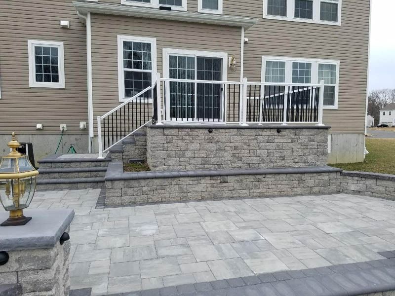 Backyard patio with stone pavers and retaining walls, leading to a raised deck with railings, next to a tan house.