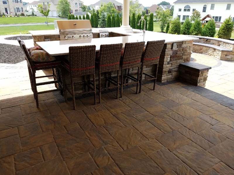 Outdoor kitchen with stone bar, seating, and grill on a patio with a brick-like pattern.
