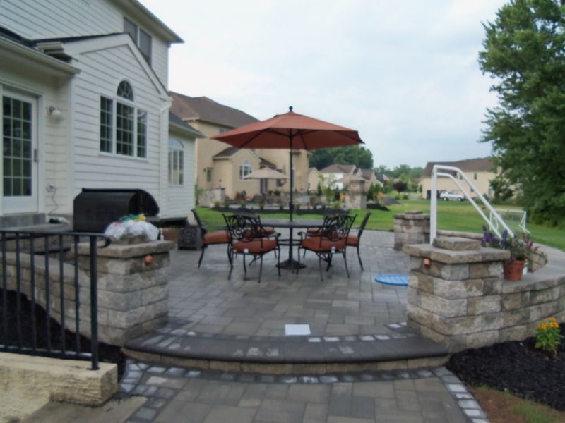 Patio with table, chairs, and umbrella outside a house; brickwork, dark grill.