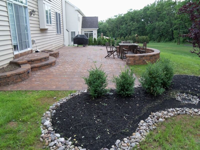 Backyard patio with brick pavers, steps, and a flower bed with mulch, shrubs, and stones.