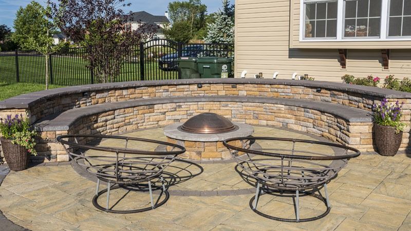 Stone fire pit with built-in seating on a patio, two metal chairs in front, near a beige house and green lawn.