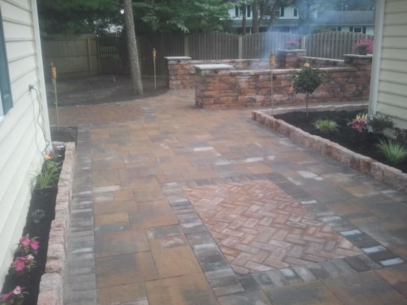 Brick patio with a brick wall and planters, trees in the background, and a light-colored house to the left.