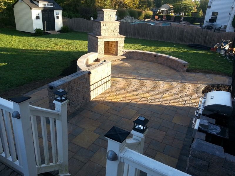 Backyard patio with fireplace, stone walls, and stamped concrete, with a deck in foreground.