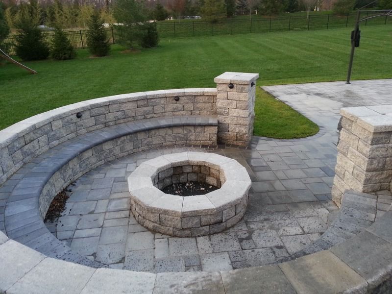 Stone fire pit surrounded by a curved stone bench on a paved patio, with a grassy lawn in the background.