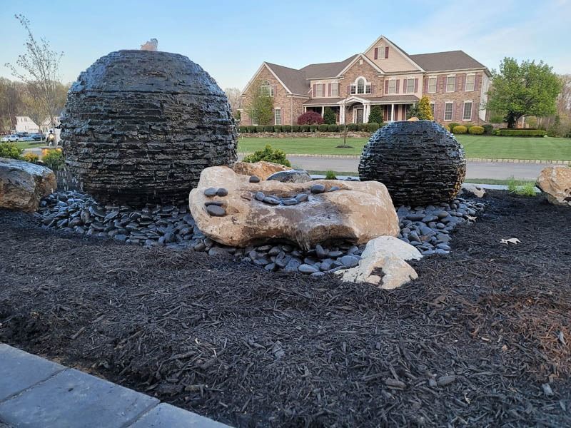 Decorative stone water fountain in front of a large house, surrounded by black mulch and stones.