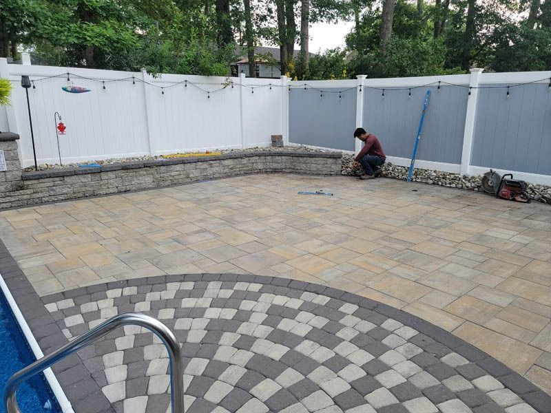 A man works on a patio with a retaining wall and fence near a pool. The patio has a unique brick design.
