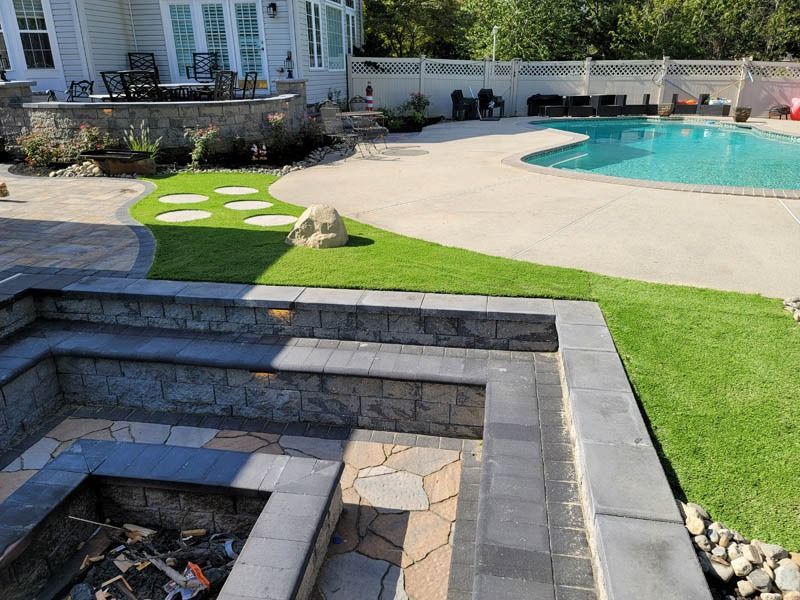 Stone fire pit and patio with pool, lawn, and house in background.
