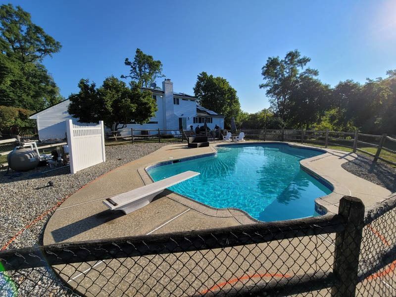 Swimming pool in backyard of white house, surrounded by trees and a fence.