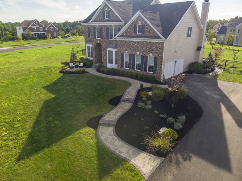 Two-story house with stone and beige facade, winding walkway, landscaped garden, and long driveway on green lawn.