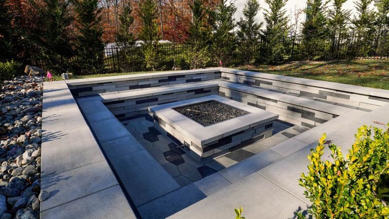 Stone fire pit with built-in seating in a backyard setting surrounded by landscaping.