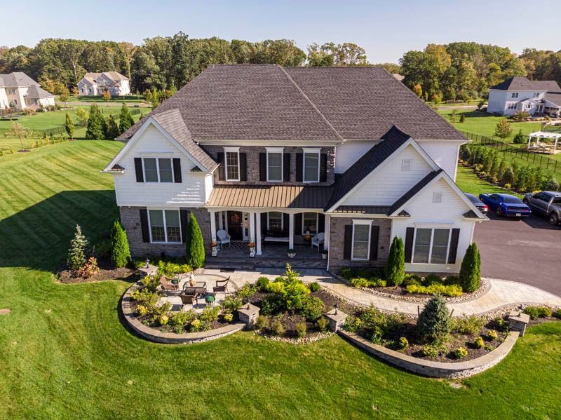 Two-story white house with dark shutters, stone accents, and landscaped front yard on a sunny day.