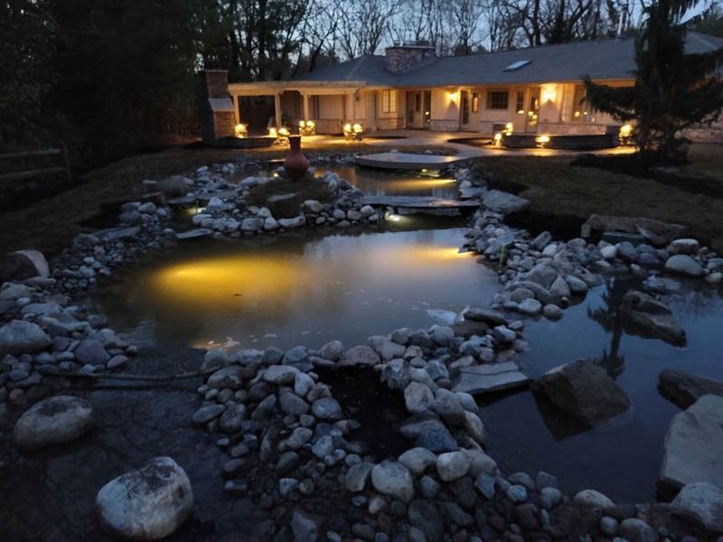 Night view of a pond with large rocks and lighting in front of a house.