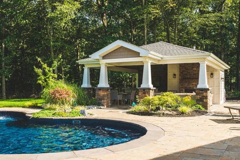 Poolside cabana with pillars, stone facade, and seating area near a pool surrounded by landscaping and trees.