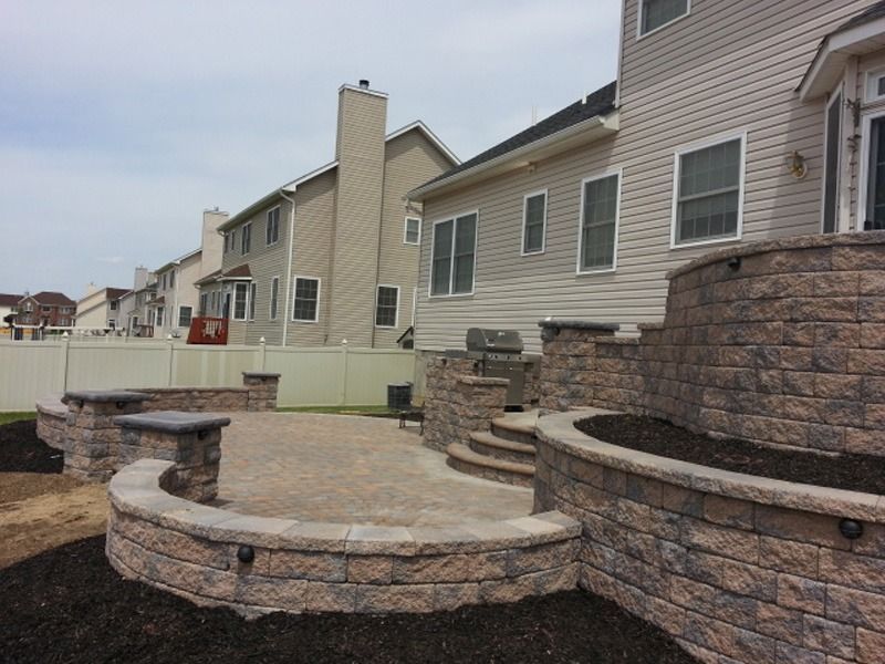 Backyard patio with tiered retaining walls, stairs, and grill. Tan brick, beige siding, and cloudy sky.